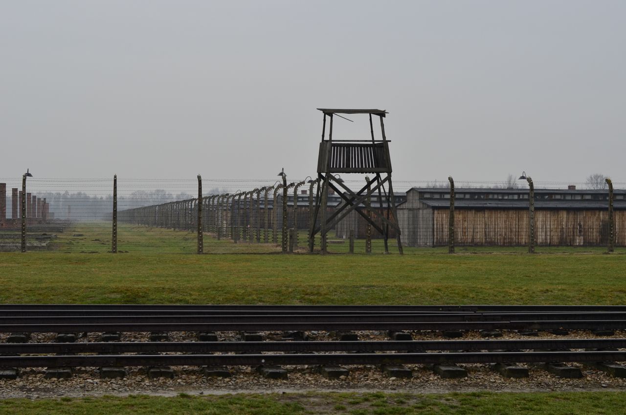 Een sombere foto van concentratiekamp Auschwitz met een spoorweg en een beveiligingstoren en hekken.
